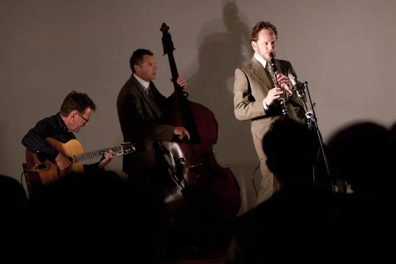 Swing 42 as a trio on stage: guitar, double bass and clarinet, their shadows on the wall behind